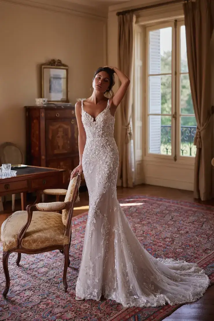 A woman stands gracefully in a beautiful wedding dress with intricate detailing, posing elegantly in a well-lit room with vintage decor and large windows.