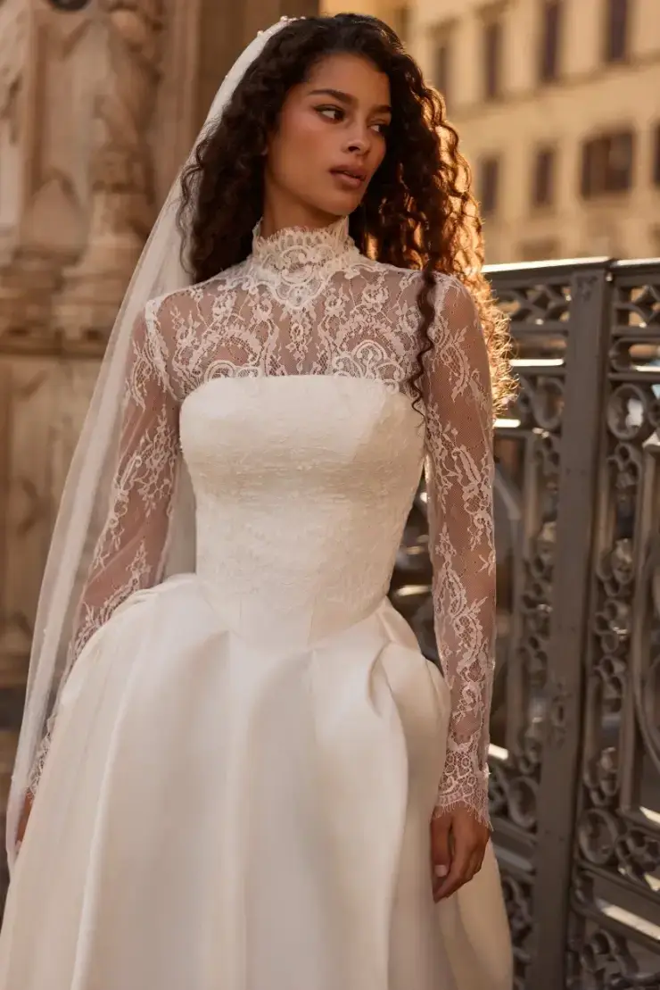 A woman in a white wedding dress with lace long sleeves and a veil, standing outdoors near ornate architecture.