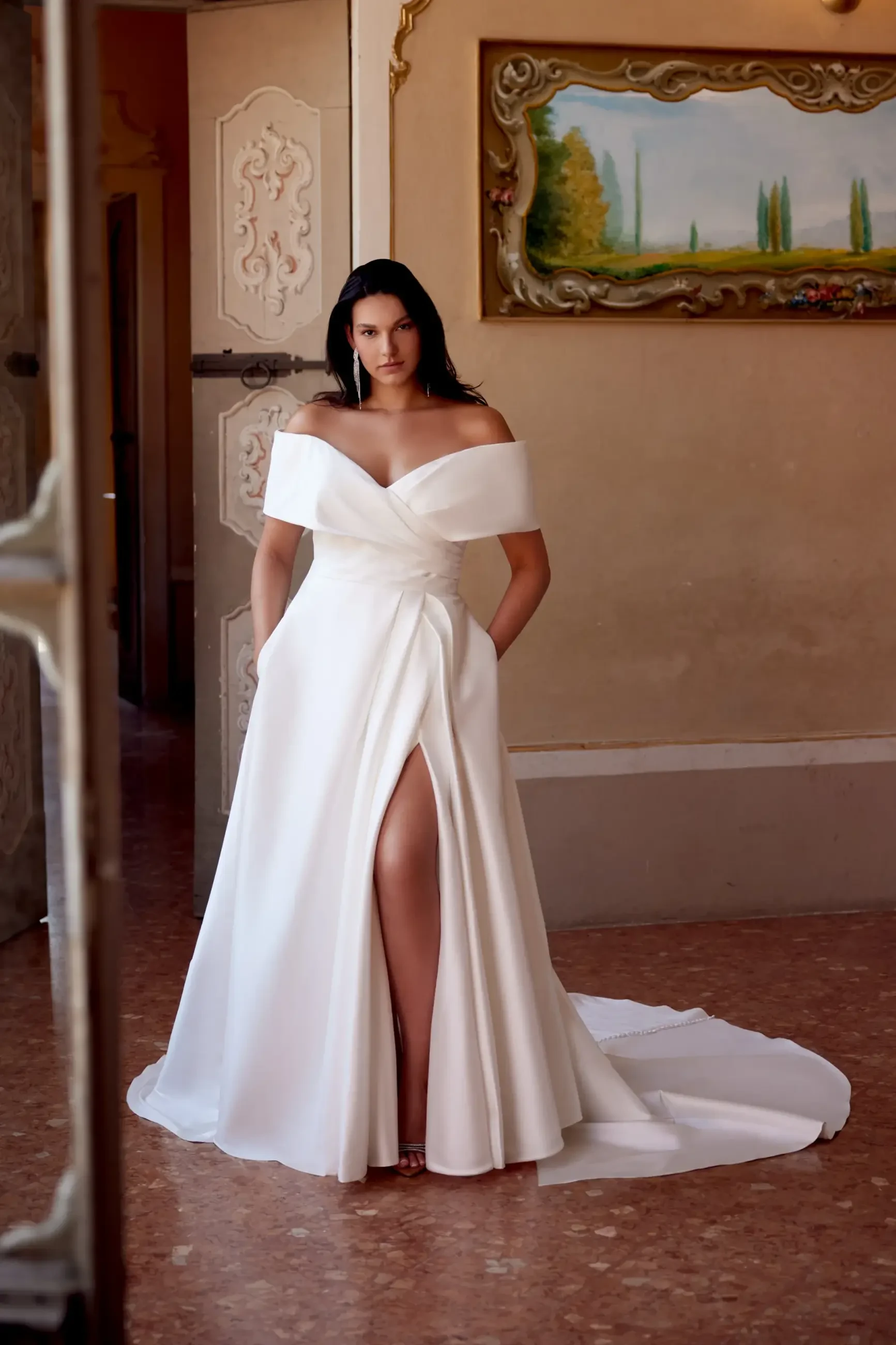 A woman in a white, off-the-shoulder gown with a high slit, standing confidently in a beautifully decorated room.