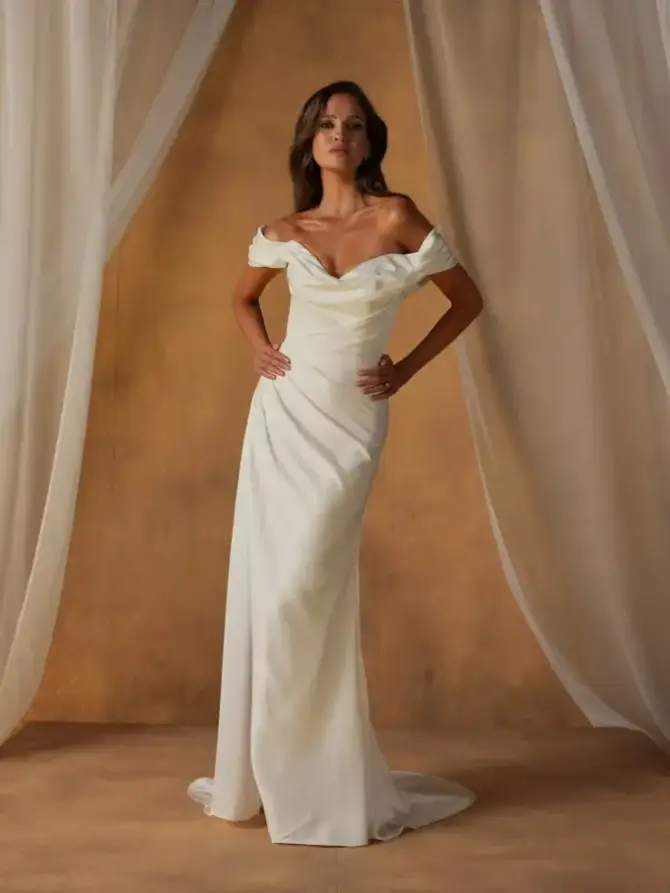 A woman in an elegant off-the-shoulder white gown poses confidently against a soft, neutral backdrop.